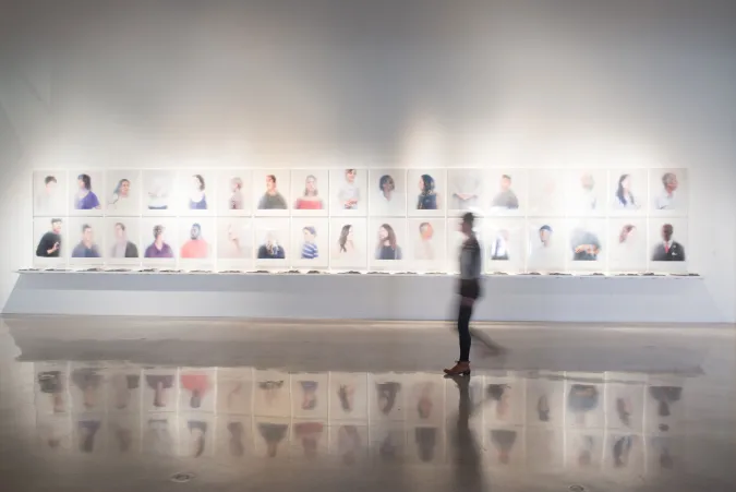 person walking in front of rows of framed portraits installed on wall in gallery