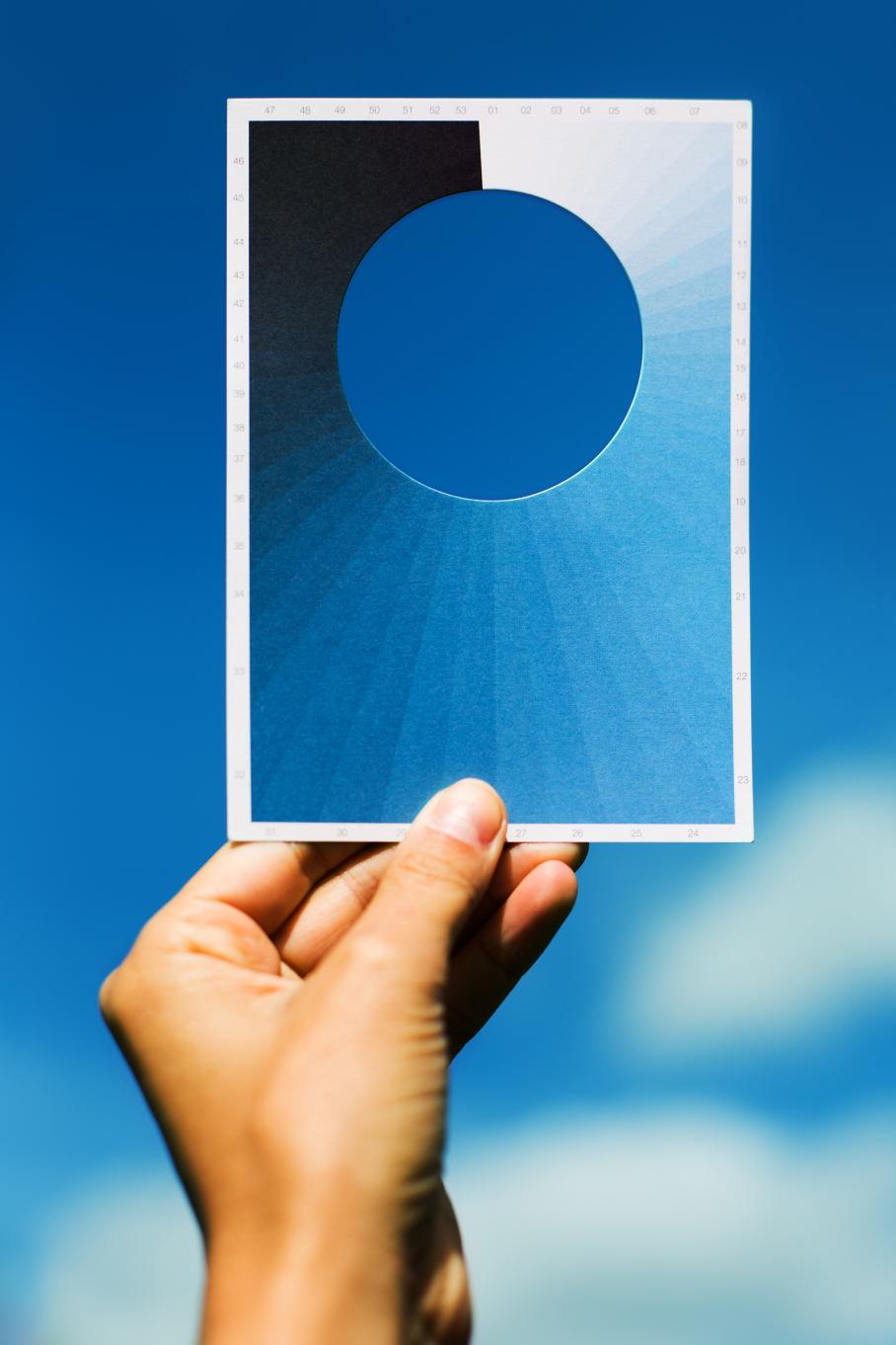 A hand holding a postcard against a blue sky. The postcard contains a spectrum of blue hues and has a circular die cut near the top.