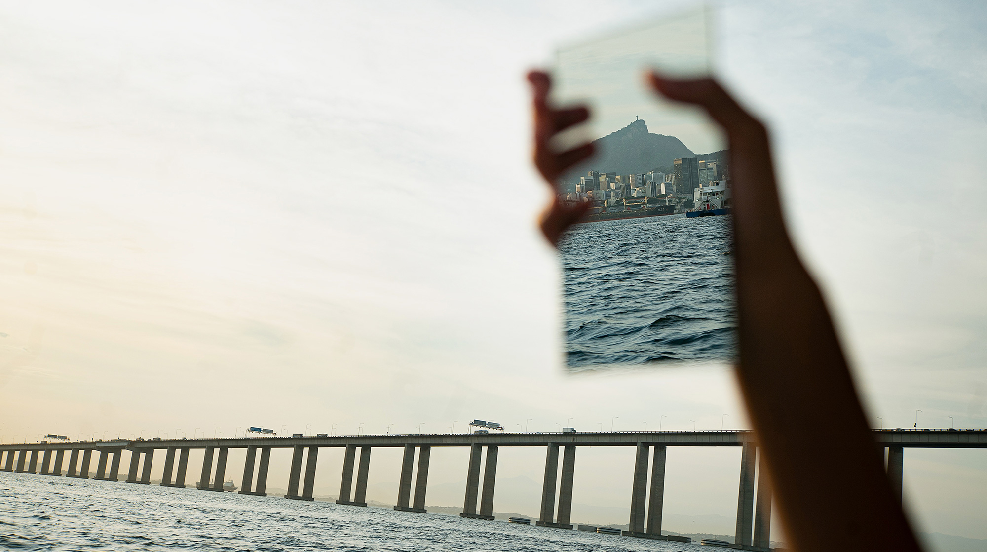 hand holding mirror that's reflecting mountains, with bridge crossing open water in the background