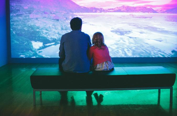 adult and child sitting on bench watching video installation